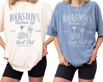 May include: Two t-shirts, one cream and one blue, each with the text "BOOKS DOWN Bottoms Up Book Club EDMONDS WA EST 2006" in a navy blue design. The shirts are short-sleeved and worn with black shorts.
