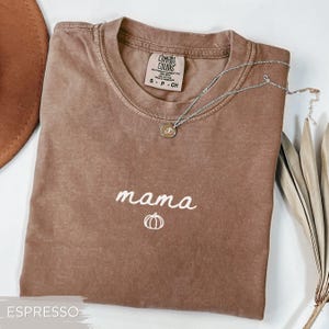 May include: Espresso-coloured t-shirt with the word "mama" in white script and a pumpkin graphic. A silver necklace with a pendant rests on the shirt. The shirt is folded on a white surface.