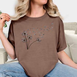 May include: A brown t-shirt with a graphic of a dandelion blowing in the wind. The dandelion is made up of small circles that resemble seeds.