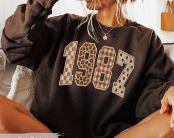 May include: A dark chocolate brown long-sleeve sweatshirt featuring the number 1987 printed across the chest. Each digit has a unique pattern: the 1 is checkered, the 9 is leopard print, the 8 is plaid, and the 7 is polka-dotted.