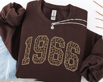 May include: A dark chocolate-colored sweatshirt with the year "1966" in a leopard print design. A silver necklace with a pendant is draped across the sweatshirt. The words "DARK CHOCOLATE" are at the bottom of the image.