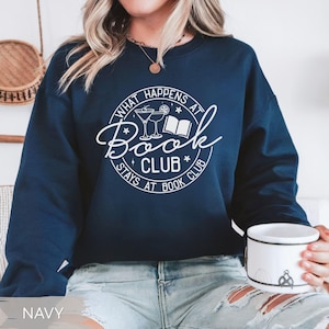 May include: Navy blue sweatshirt with the text "What Happens at Book Club Stays at Book Club" in a white circular design. The design includes illustrations of cocktails and a book. The sweatshirt is a crew neck style.