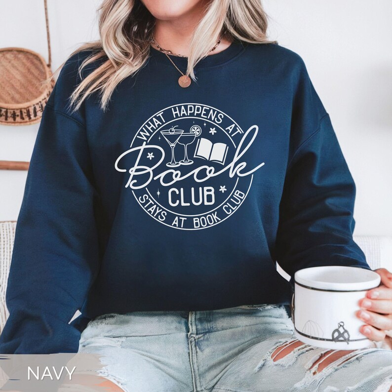 May include: Navy blue sweatshirt with the text "What Happens at Book Club Stays at Book Club" in a white circular design. The design includes illustrations of cocktails and a book. The sweatshirt is a crew neck style.
