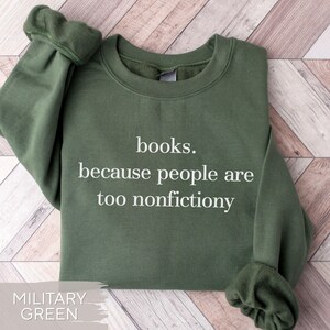 May include: Military green sweatshirt with the text "books. because people are too nonfictiony" in white. The sweatshirt is a crew neck style with long sleeves. The background is a light wood pattern.