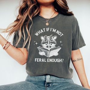May include: A gray t-shirt with a white graphic of a raccoon reading a book. The text on the shirt reads "What if I'm not feral enough?"