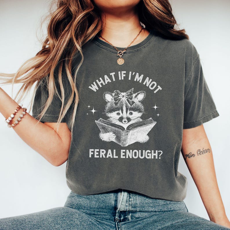 May include: A gray t-shirt with a white graphic of a raccoon reading a book. The text on the shirt reads "What if I'm not feral enough?"