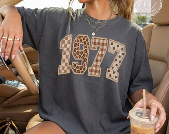 May include: A charcoal gray short-sleeve t-shirt featuring the number 1972 printed in large block letters. Each digit has a distinct pattern: a checkerboard, leopard print, brown plaid, and polka dots. The shirt has a crew neckline.