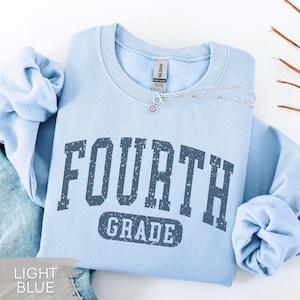 May include: A light blue crewneck sweatshirt featuring the text FOURTH GRADE in a dark blue, distressed, collegiate-style font. The garment includes a Gildan brand label at the inner neckline and a silver-toned chain necklace resting on the collar.