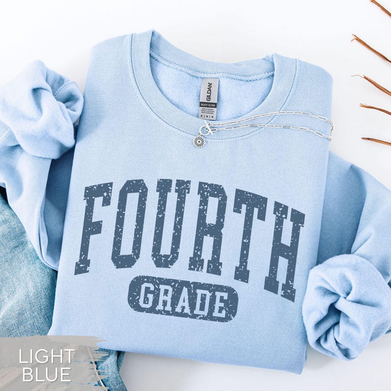 May include: A light blue crewneck sweatshirt featuring the text FOURTH GRADE in a dark blue, distressed, collegiate-style font. The garment includes a Gildan brand label at the inner neckline and a silver-toned chain necklace resting on the collar.