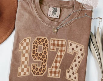 May include: A brown short-sleeve t-shirt featuring the number 1972 printed in large block letters. Each digit has a unique pattern: a checkerboard, leopard print, gingham plaid, and polka dots. A silver necklace rests on the neckline.