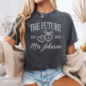 May include: A dark gray t-shirt with the text "THE FUTURE" above "EST 2026" and "Mrs. Johnson" in white script. Two heart-shaped pendants with the letters "T" and "S" are in the center. The shirt is worn by a person.