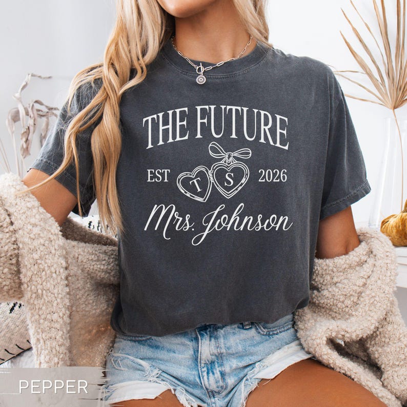 May include: A dark gray t-shirt with the text "THE FUTURE" above "EST 2026" and "Mrs. Johnson" in white script. Two heart-shaped pendants with the letters "T" and "S" are in the center. The shirt is worn by a person.