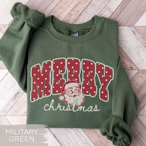 May include: A military green crewneck sweatshirt featuring the word MERRY in large red letters with white polka dots. Below the text is a graphic of a Santa Claus face and the word christmas written in white cursive script.