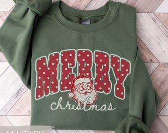 May include: A military green crewneck sweatshirt featuring the word MERRY in large red letters with white polka dots. Below the text is a graphic of a Santa Claus face and the word christmas written in white cursive script.