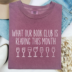 May include: A mauve colored t-shirt with the text "What our book club is reading this month" and a row of seven white outline drawings of wine glasses.