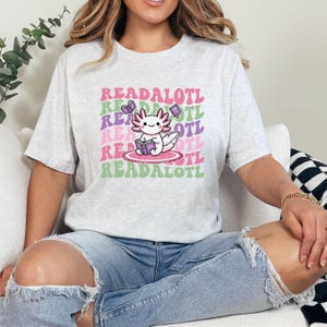 May include: A gray t-shirt with a pink and green graphic of an axolotl reading a book. The text "READALOTL" is repeated in a retro font.