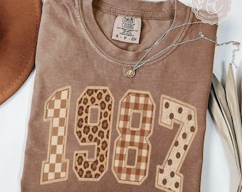 May include: A brown crewneck t-shirt featuring the year 1987 in large block letters. Each digit has a unique pattern: the 1 is checkered, the 9 is leopard print, the 8 is gingham, and the 7 is polka-dotted. A silver necklace rests on the neckline.