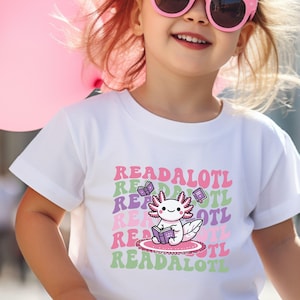 May include: White t-shirt with a pink axolotl wearing glasses and reading a book. The text "READALOTL" is repeated in a retro font in pink, green, and purple.