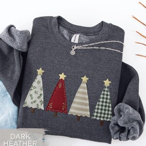 May include: Dark heather gray sweatshirt with four appliqued Christmas trees in various patterns and colors, topped with yellow stars. A silver necklace with a sun pendant is draped across the sweatshirt. The text "DARK HEATHER" is at the bottom.