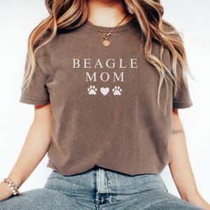 K&ouml;nnte beinhalten: Ein braunes T-Shirt mit dem Text "BEAGLE MOM" in wei&szlig;en Buchstaben und zwei wei&szlig;en Pfotenabdr&uuml;cken auf beiden Seiten eines wei&szlig;en Herzens.