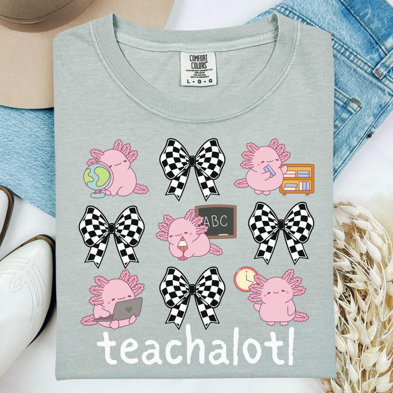 May include: A light grey Comfort Colors t-shirt with a repeating pattern of pink axolotls with school-themed accessories and black and white checkered bows. The word "teachalotl" is printed at the bottom.