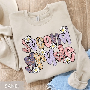 May include: Sand-colored sweatshirt with the words "SECOND GRADE" in a colorful, floral design. The letters are filled with patterns and decorated with white daisy flowers. The sweatshirt is folded on a light-colored surface.