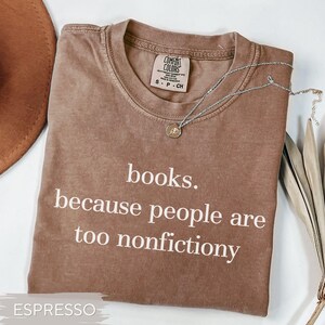 May include: Espresso-colored t-shirt with the text "books. because people are too nonfictiony" in white. The shirt has a crew neck and short sleeves. A silver necklace with a small pendant is draped on the shirt.