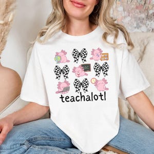 May include: White t-shirt with a repeating pattern of pink axolotls with school-themed accessories and black and white checkered bows. The text "teachalotl" is printed in black at the bottom of the shirt.