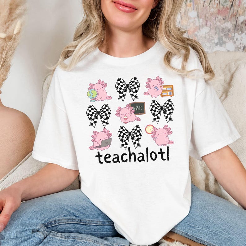 May include: White t-shirt with a repeating pattern of pink axolotls with school-themed accessories and black and white checkered bows. The text "teachalotl" is printed in black at the bottom of the shirt.
