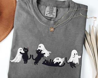 May include: A gray t-shirt with a graphic of black cats interacting with white ghosts. The shirt has a crew neck and short sleeves. A gold necklace with a pendant is draped across the shirt. The shirt is labeled "Comfort Colors".