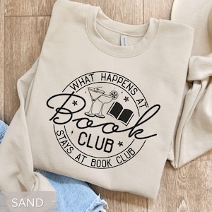 May include: Sand-colored sweatshirt with a black graphic that reads "What Happens at Book Club Stays at Book Club." The design includes cocktail glasses and an open book. The sweatshirt is a crew neck style.