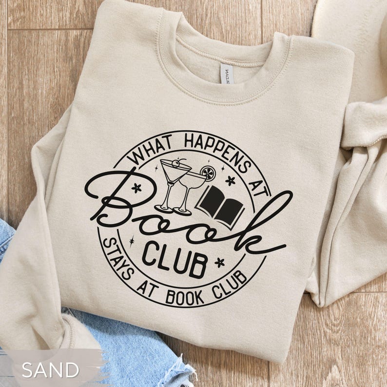 May include: Sand-colored sweatshirt with a black graphic that reads "What Happens at Book Club Stays at Book Club." The design includes cocktail glasses and an open book. The sweatshirt is a crew neck style.