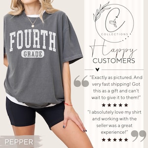 May include: A charcoal gray short-sleeve t-shirt featuring the white distressed text "FOURTH GRADE" on the chest. The shirt is worn over a white undershirt and paired with black shorts. The image includes a logo and customer review text.