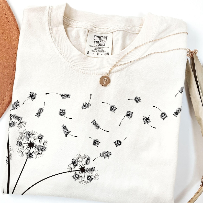 May include: A white t-shirt with a black graphic of raccoons blowing dandelion seeds. The shirt has a tag that says "Comfort Colors" and the size "S - P - CH".