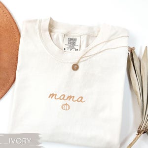 May include: An ivory t-shirt featuring the word "mama" and a pumpkin graphic in brown. A gold necklace with a circular pendant lies on the shirt. The shirt is folded, with a brown hat and dried leaves in the background.