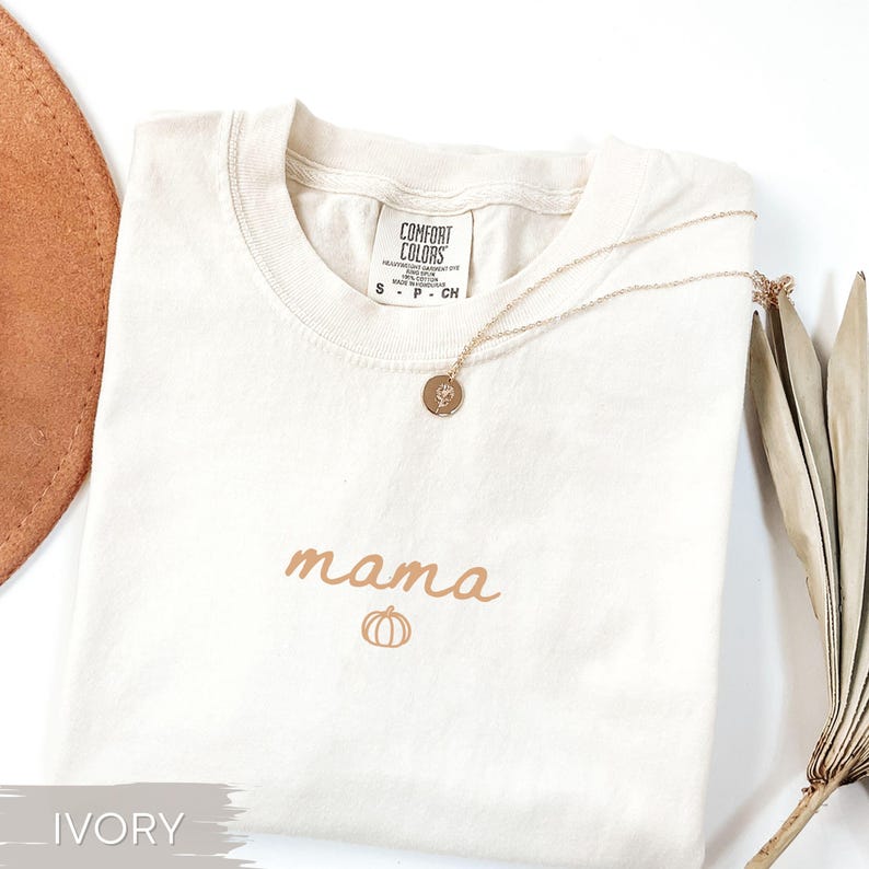 May include: An ivory t-shirt featuring the word "mama" and a pumpkin graphic in brown. A gold necklace with a circular pendant lies on the shirt. The shirt is folded, with a brown hat and dried leaves in the background.