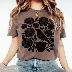 May include: Espresso-colored t-shirt featuring a black floral and bird graphic. The design includes a raven perched on a coffee cup, surrounded by peony-like flowers. The shirt is a short-sleeved crew neck.