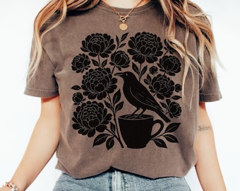 May include: Espresso-colored t-shirt featuring a black floral and bird graphic. The design includes a raven perched on a coffee cup, surrounded by peony-like flowers. The shirt is a short-sleeved crew neck.