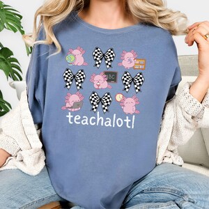 May include: A blue t-shirt with a repeating pattern of pink axolotls, black and white checkered bows, books, and a chalkboard with "ABC". The text "teachalotl" is printed below the axolotl design.