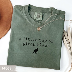 May include: Moss green t-shirt with the text "a little ray of pitch black" and a black crow graphic. A gold-colored necklace with a pendant rests on the shirt. The shirt is folded and has a Comfort Colors tag.