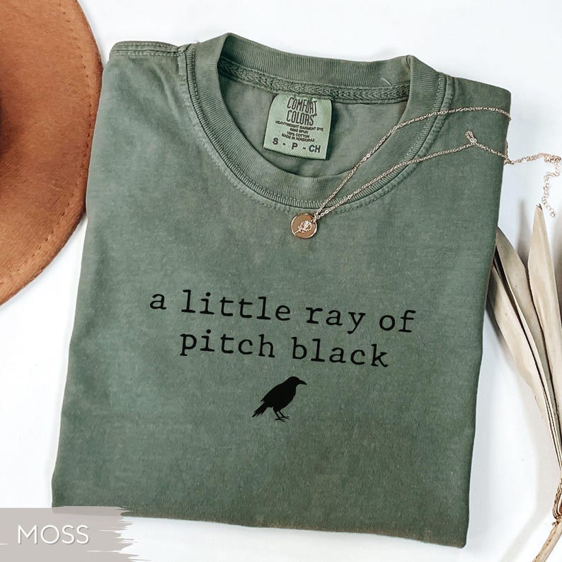 May include: Moss green t-shirt with the text "a little ray of pitch black" and a black crow graphic. A gold-colored necklace with a pendant rests on the shirt. The shirt is folded and has a Comfort Colors tag.