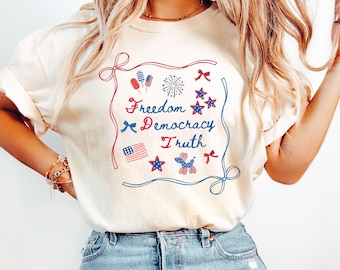 May include: Cream-coloured t-shirt with a patriotic design in red and blue, featuring the words "Freedom Democracy Truth". The design includes stars, bows, an American flag, and fireworks. The shirt is a casual style.