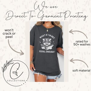 May include: A dark gray t-shirt with a white graphic of a raccoon reading a book. The text on the shirt reads "What if I'm not feral enough?"
