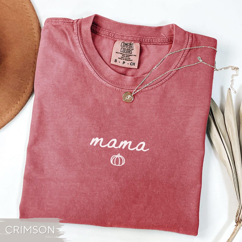 May include: Crimson-coloured t-shirt with the word "mama" and a pumpkin graphic embroidered in white. A gold necklace with a pendant rests on the shirt. The shirt is a Comfort Colors brand, size small.