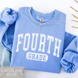 May include: A light blue crewneck sweatshirt featuring the white distressed text FOURTH GRADE printed on the front. The garment has long sleeves with ribbed cuffs and a ribbed collar. A silver chain necklace with a small circular pendant rests on the neckline.