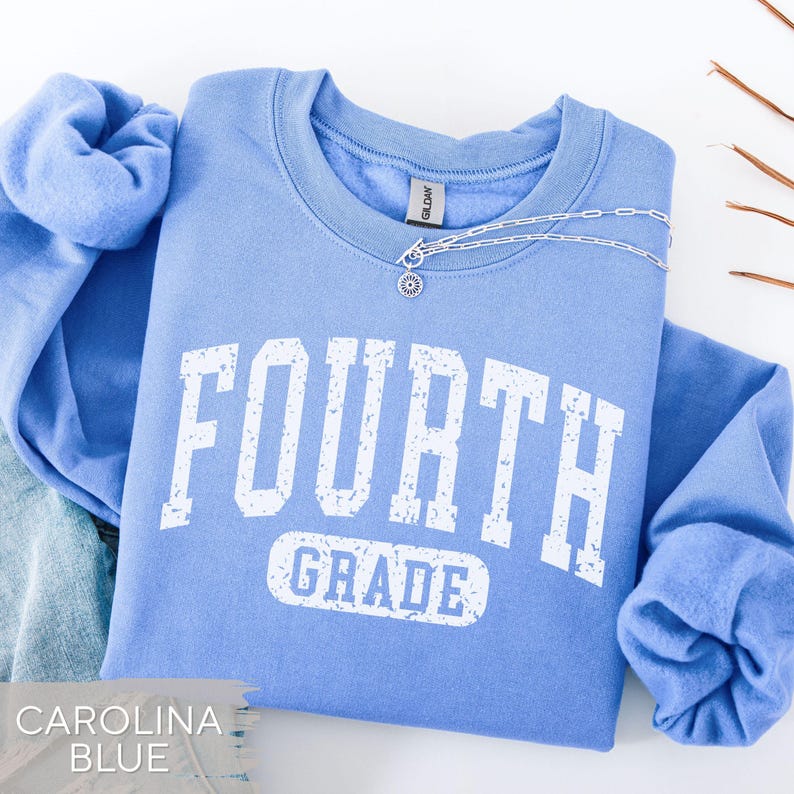 May include: A light blue crewneck sweatshirt featuring the white distressed text FOURTH GRADE printed on the front. The garment has long sleeves with ribbed cuffs and a ribbed collar. A silver chain necklace with a small circular pendant rests on the neckline.