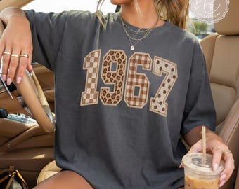 May include: A charcoal gray short-sleeve t-shirt featuring the number 1967 printed in large block letters. Each digit has a unique pattern: the 1 is checkered, the 9 is leopard print, the 6 is plaid, and the 7 is polka-dotted in brown tones.