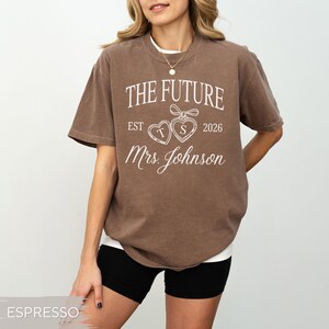 May include: Espresso-colored t-shirt with the text "THE FUTURE" above "EST 2026 Mrs. Johnson". The design includes two heart-shaped lockets. The shirt is a relaxed fit and has short sleeves.