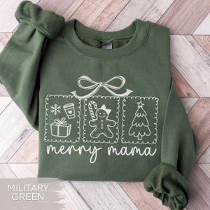 May include: Military green sweatshirt with a white graphic including a bow, snowflake, coffee cup, gift, gingerbread person, and Christmas tree. The words "merry mama" are written below the graphic. The sweatshirt is folded on a light wood-grain surface.