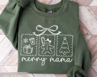 May include: Military green sweatshirt with a white graphic featuring a bow, snowflake, coffee cup, gift, gingerbread person, and Christmas tree. The words "merry mama" are written below the graphic. The sweatshirt is folded on a light wood-grain surface.